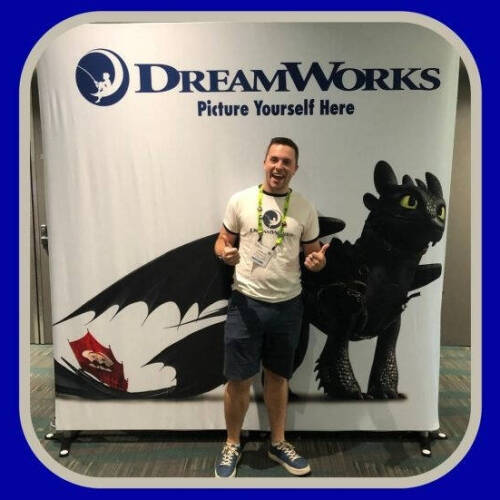 A person is standing in front of a DreamWorks promotional backdrop featuring Toothless, the dragon from "How to Train Your Dragon." The backdrop has the DreamWorks logo and the phrase "Picture Yourself Here." The person is giving two thumbs up and wearing